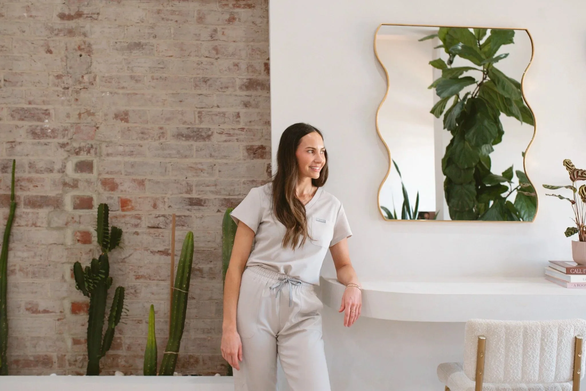 Jessica Stacey, founder of Forme Cosmetic Science, A woman with long brown hair, wearing light-colored medical scrubs, standing in a modern indoor space with plants, a mirror, and a brick wall.