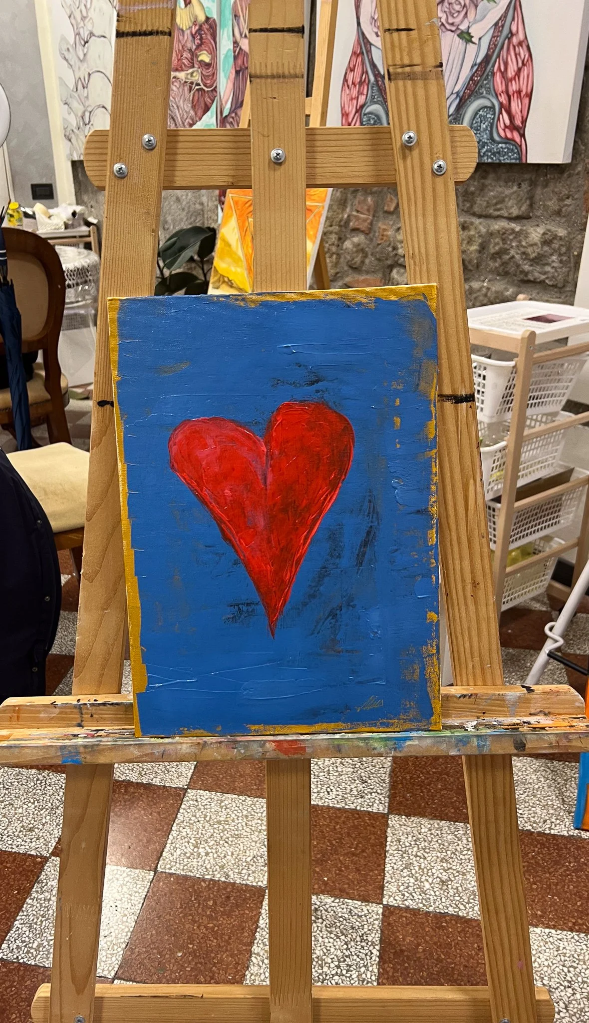 A small painting of a red heart on a blue background, displayed on a wooden easel in an art studio with paintings and art supplies in the background.