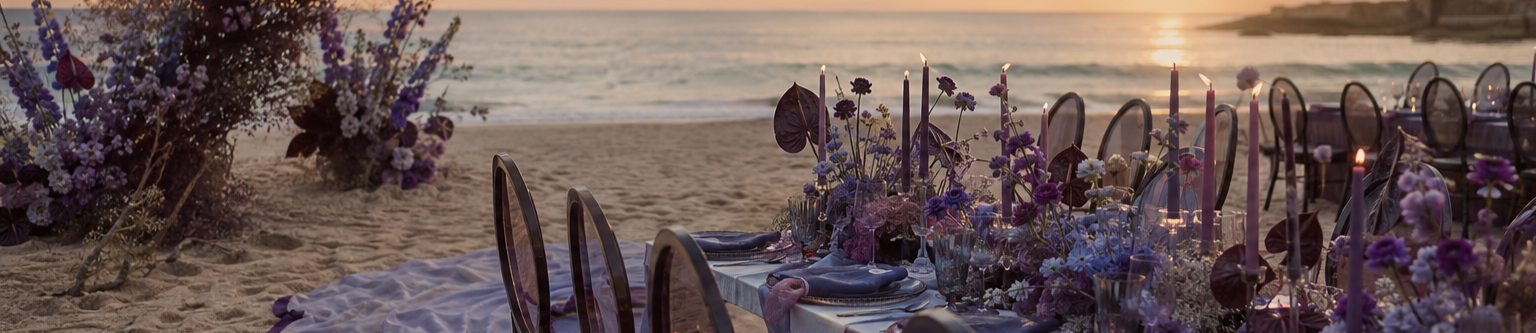 Beachside dining setup with a long table decorated with purple and white flowers, lit candles, and chairs, overlooking the ocean at sunset.