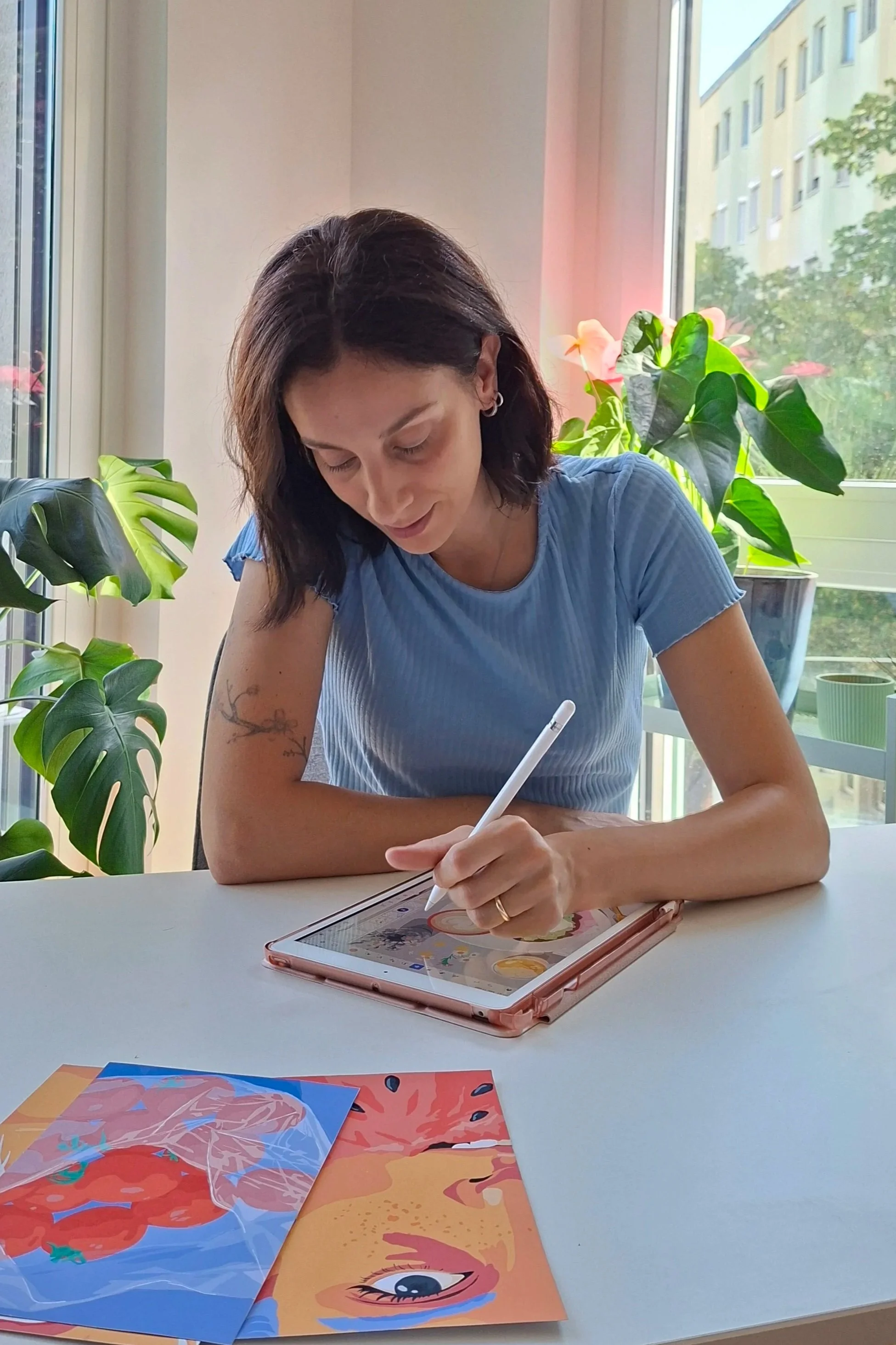 A woman with short dark hair and earrings sitting at a white table, drawing on a tablet with a white stylus. She is wearing a light blue shirt. There are green plants and a window in the background with natural light coming through.