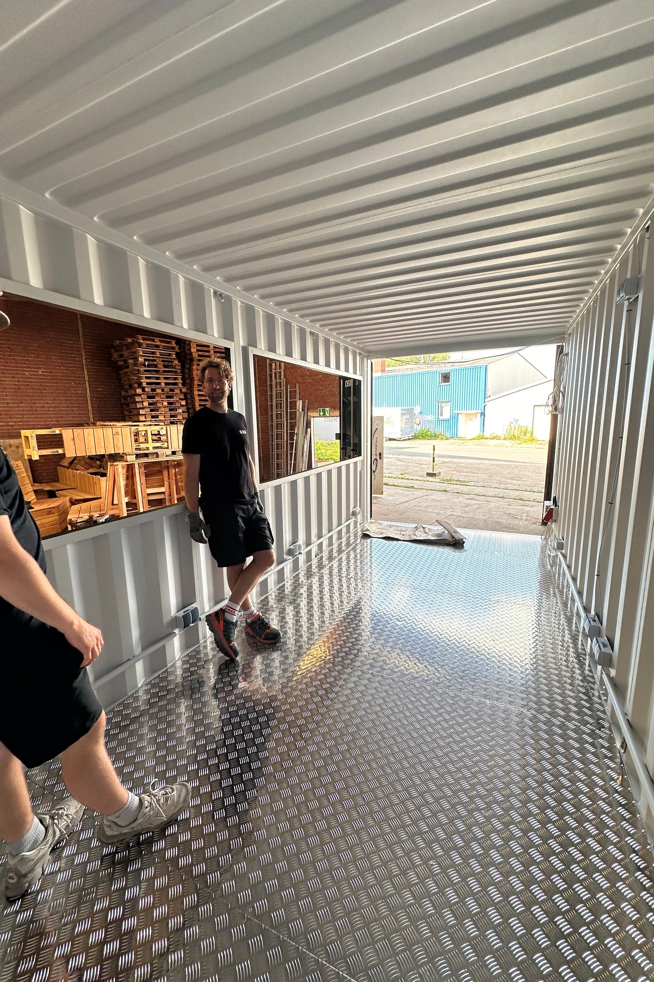 Ein Mann steht in einem modernen Container mit glänzendem Metallboden und weißen Wänden, im Hintergrund sind Holzpaletten und Gebäude sichtbar.