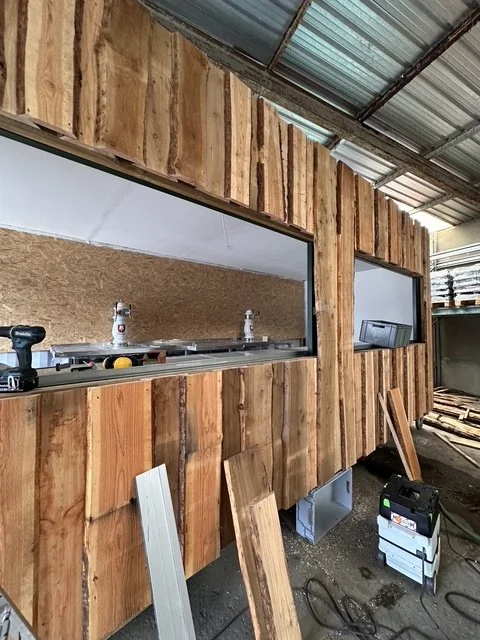 Holzwand wird in einem Bauprojekt installiert, mit Öffnungen für Fenster. Bauwerk befindet sich in einem Werkstatt- oder Lagerraum.