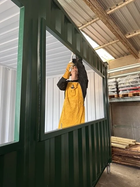 Person in gelber Arbeitsjacke schraubt an einer grünen Metallrahmenkonstruktion in einer Werkstatt.