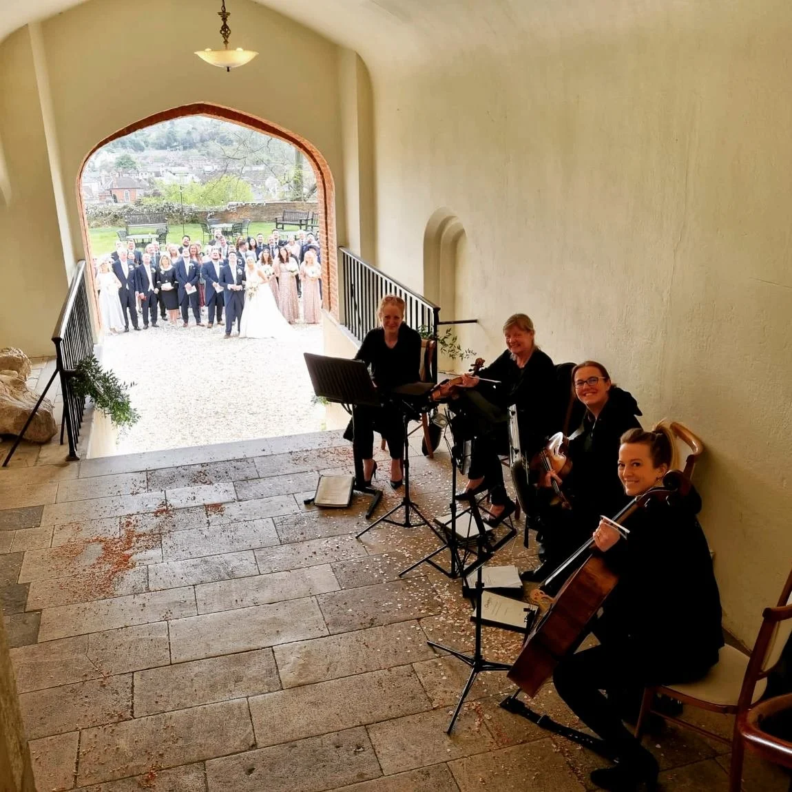A group of musicians playing string instruments inside a building, with a wedding party outside in the background.
