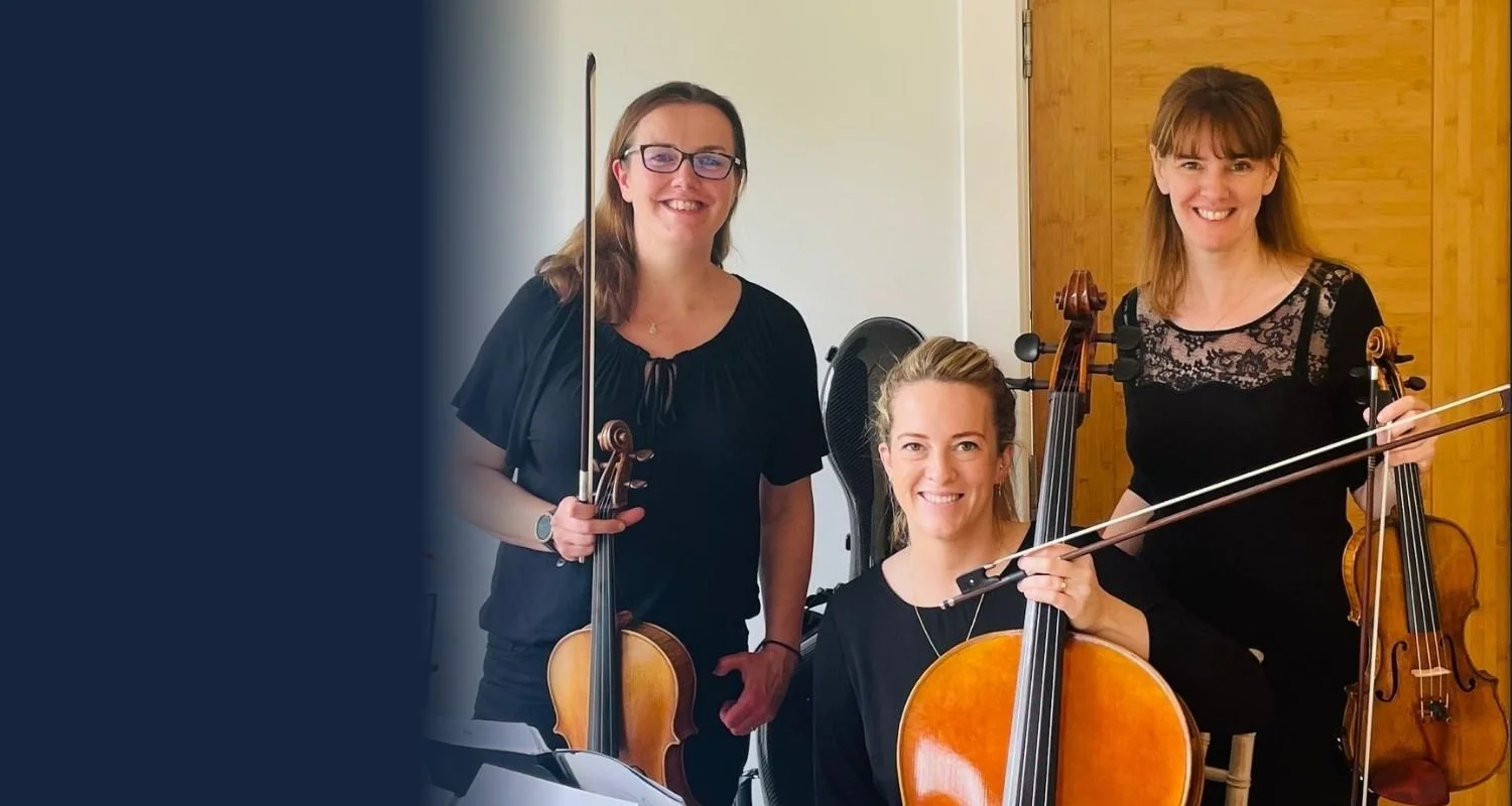 Three women holding string instruments indoors, two with violins and one with a cello, smiling at the camera.