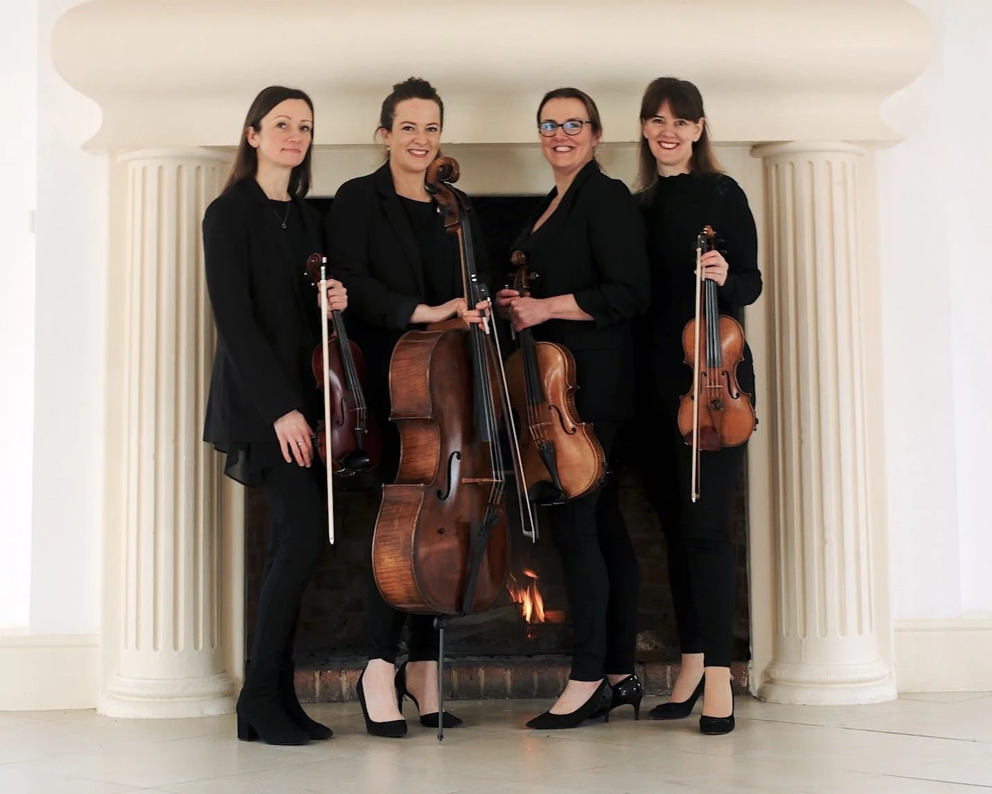 Four women dressed in black holding string instruments, standing in front of a fireplace with a fire, in a room with white walls and columns.