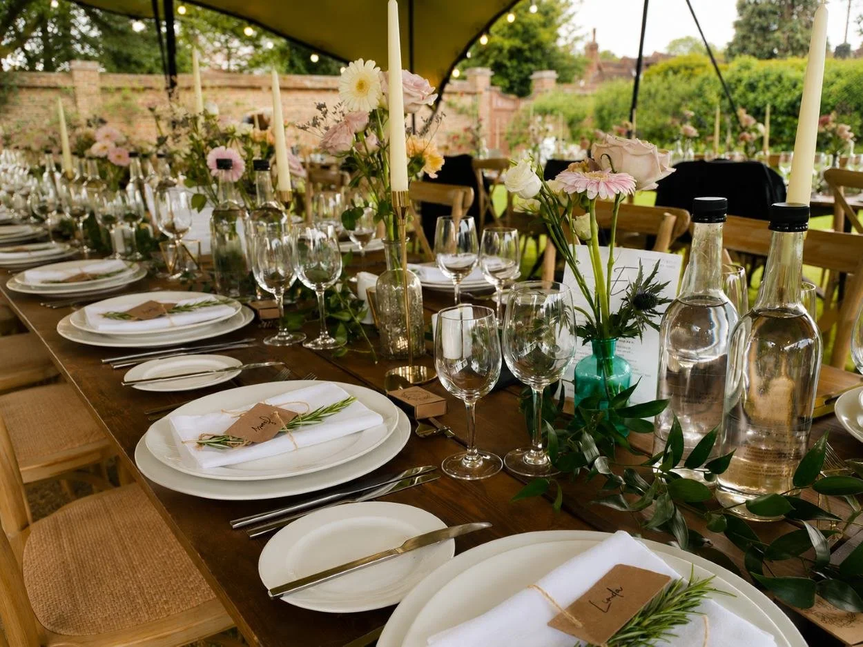 Table setup with white plates, silverware, wine glasses, candles, and floral centerpieces under a tent at an outdoor event.