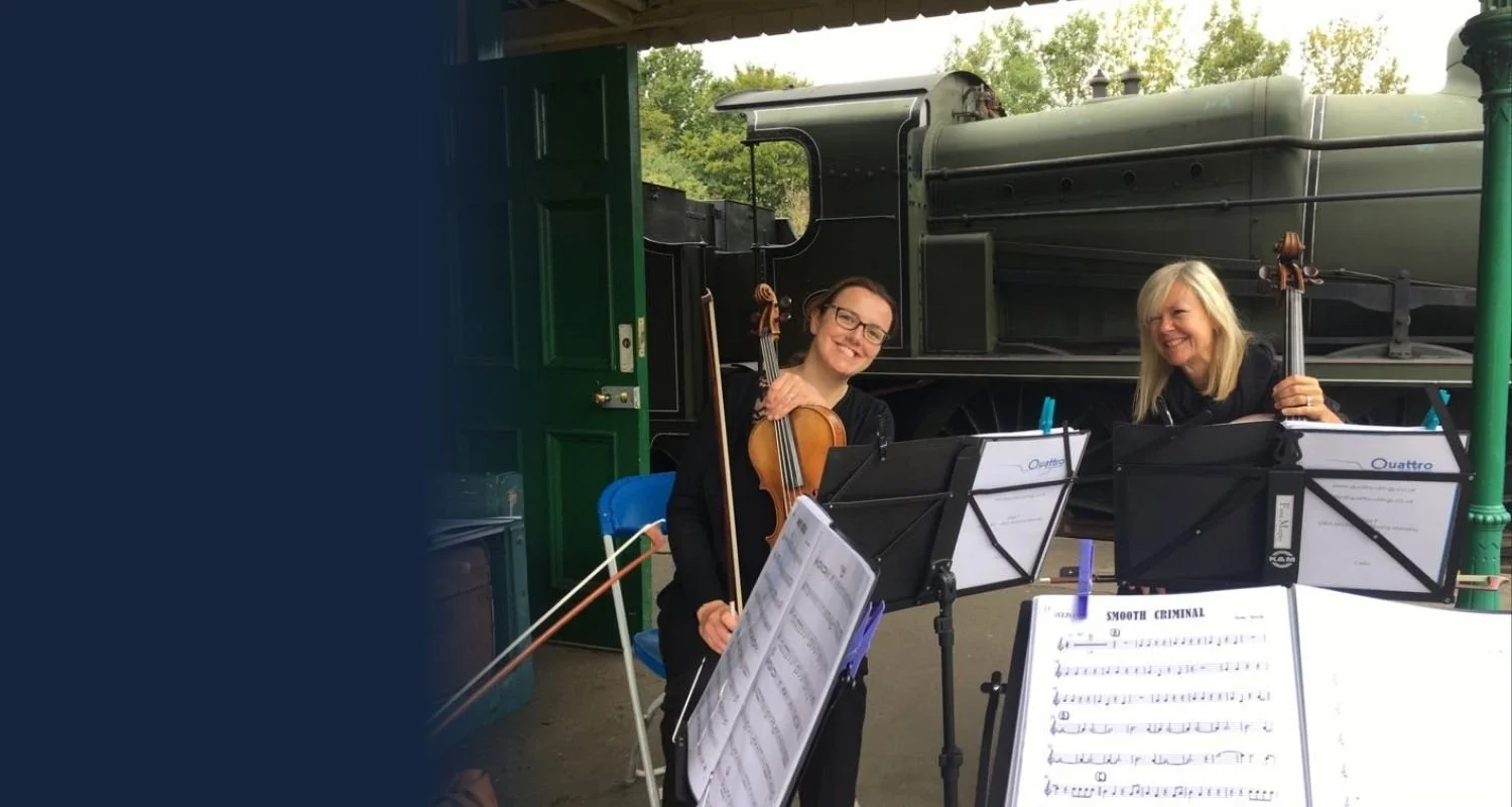 Two women with violins sitting in front of a green train engine, with sheet music in front of them, smiling and looking at the camera.