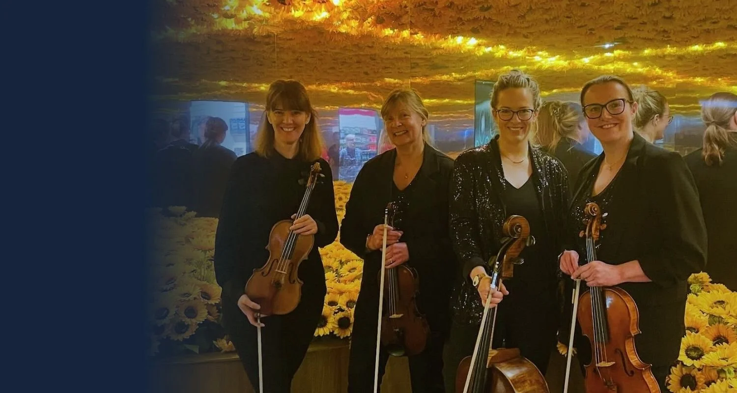 Four women standing with violins and a viola, smiling, indoors with a sunflower backdrop and ceiling resembling a sunset sky.