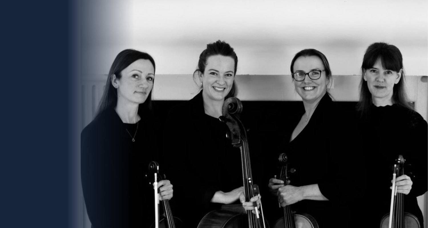 Four women standing side by side holding violins, smiling, indoors.