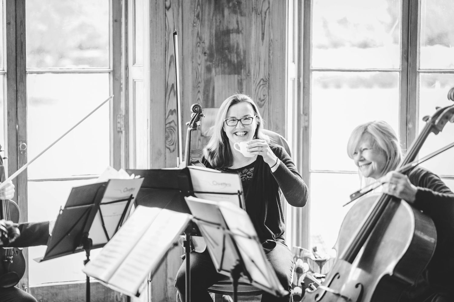 Two women in a music rehearsal space, one smiling and drinking from a cup, the other playing the cello, with sheet music on stands, sunlight coming through large windows in the background.