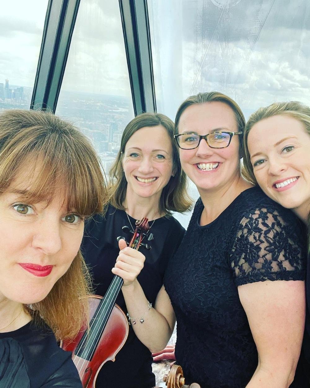 Four women smiling for a selfie in a high-rise building with city views, one holding a violin.