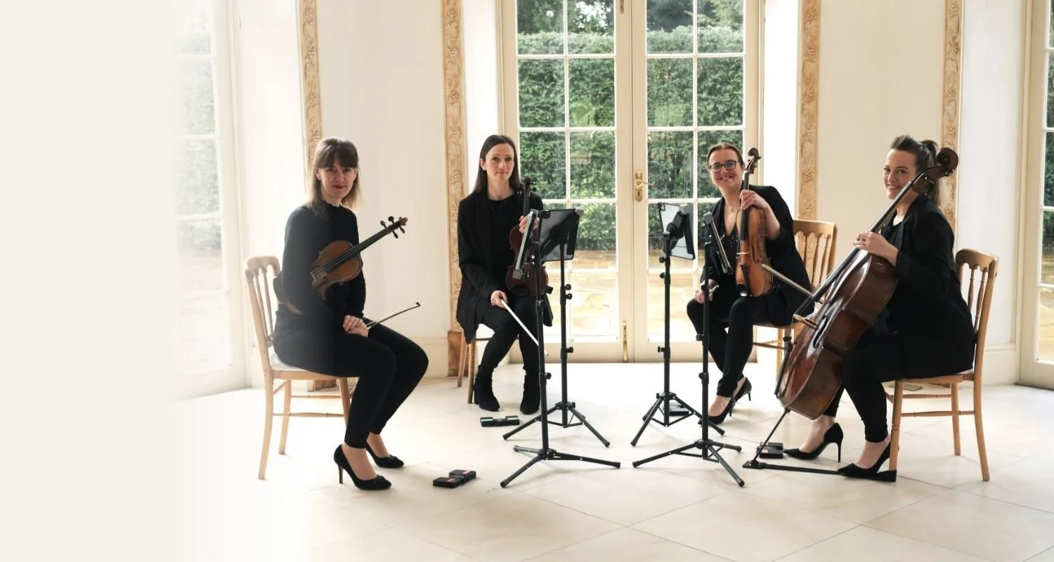 Four women in black attire sitting with string instruments in a bright room with large windows and greenery outside, performing as an orchestra.