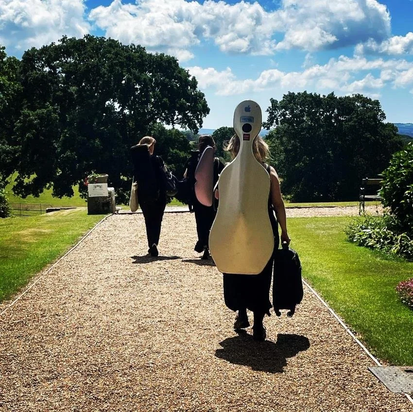 People walking on a gravel path, with one person carrying a guitar case on their back.