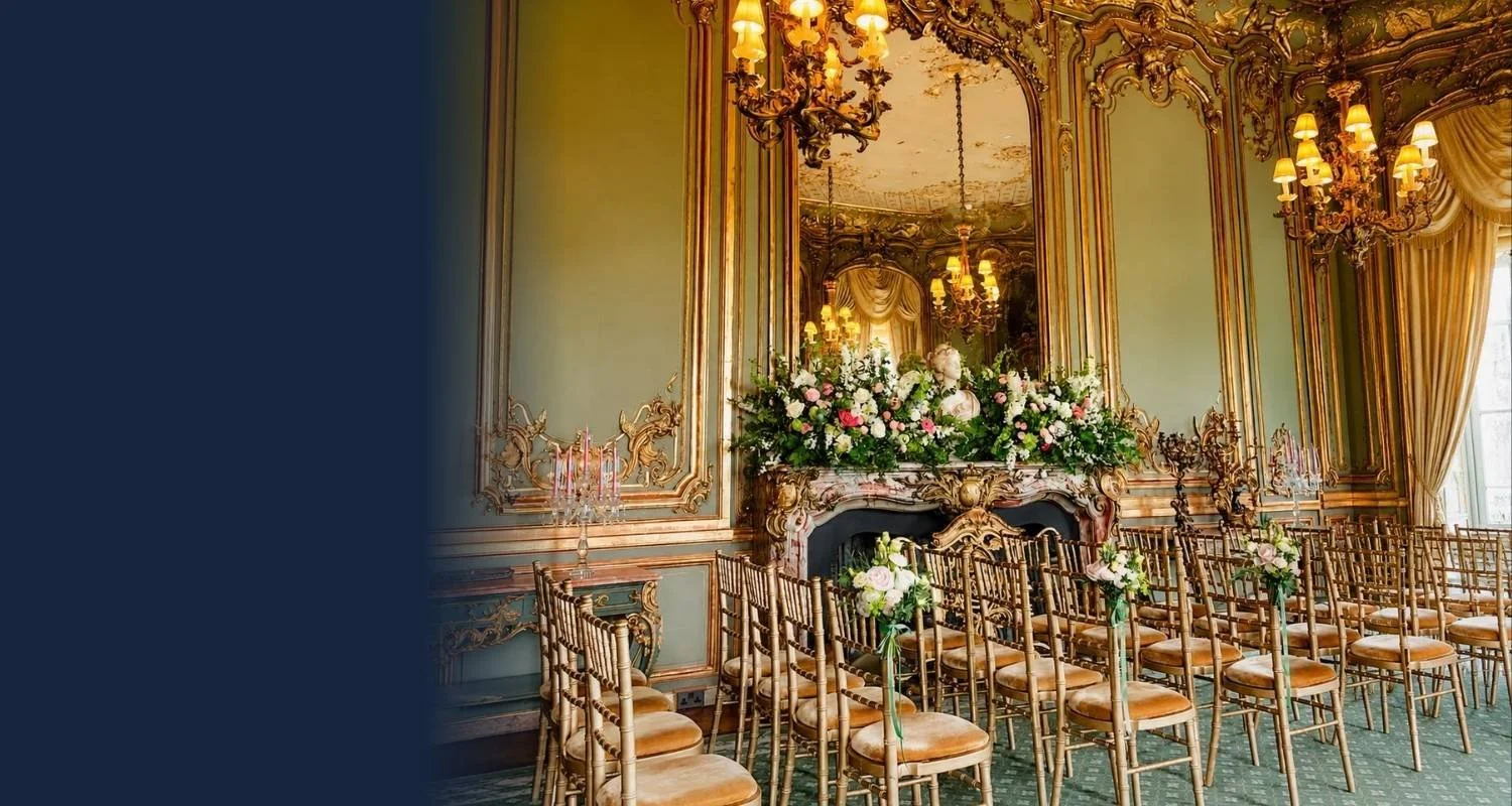 Elegant event room with ornate gold trim on pale green walls, large mirrors, chandeliers, and tall windows with drapes. Rows of chairs with floral arrangements on the aisle. Fireplace with floral decoration and a large mirror above.
