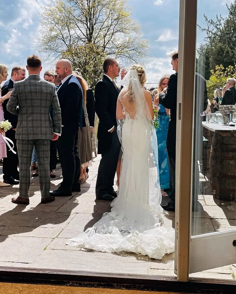 Wedding reception scene viewed from inside a building, showing a bride in a white gown and veil, a groom in a dark suit, and guests socializing outdoors on a patio under a blue sky with scattered clouds.