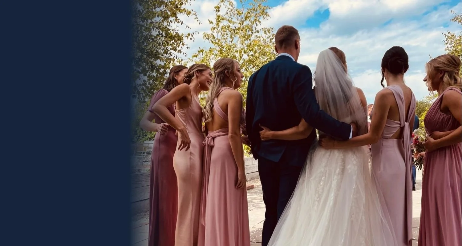A group of people at a wedding, including a bride in a white gown and veil, a groom in a navy suit, and bridesmaids in pink dresses, standing outdoors under a partly cloudy sky with trees in the background.