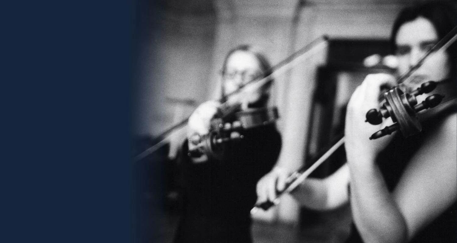 Two women playing violins together in a music practice room, both wearing glasses and focused on playing.