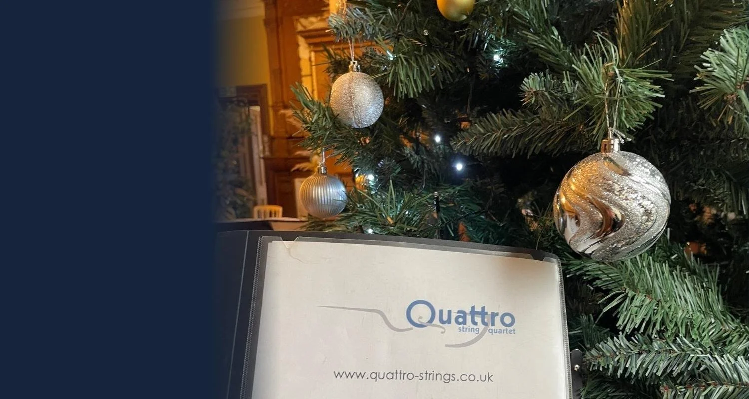 A Christmas tree decorated with silver ornaments, including a large, textured silver ball, smaller silver balls, and a string of white lights. In the foreground, there is a binder with the logo and website of Quattro Strings.