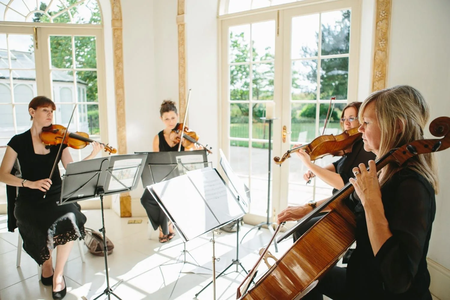 Four women playing violins and a cellist performing in a bright room with large windows showing a garden outside.