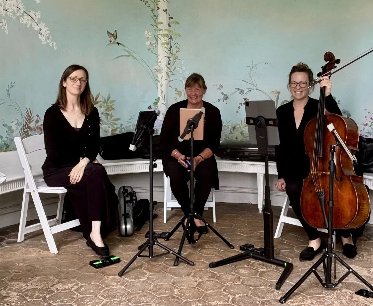 Three women, dressed in black, seated with musical instruments in a room with a pastel mural featuring birds and plants. The woman on the left is sitting next to a keyboard, the woman in the middle is on a chair with a music stand, and the woman on the right is holding a cello, with music stands and electronic devices in front of them.