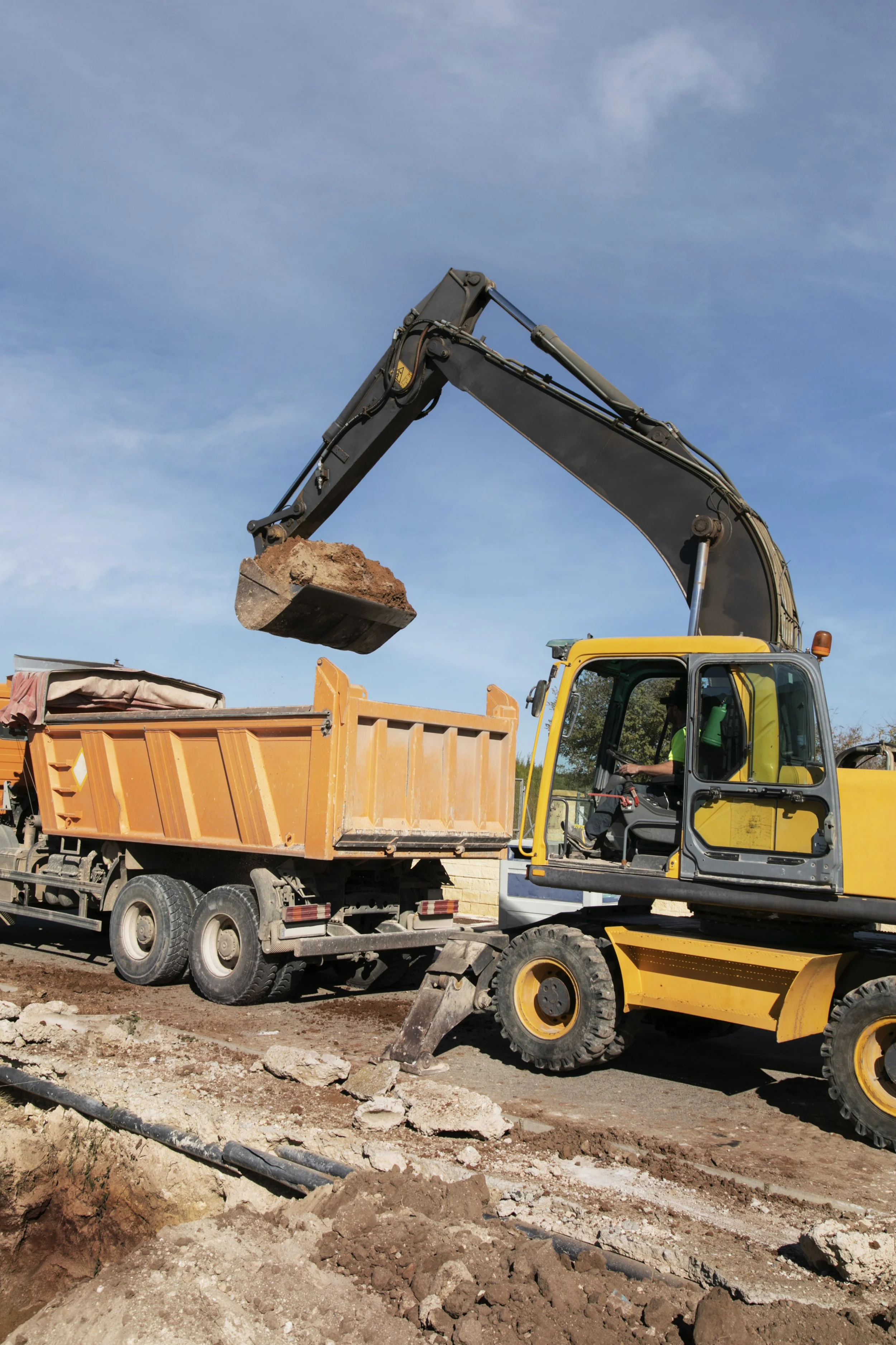 An excavator is loading dirt into a dump truck on a construction site, with a clear blue sky in the background.