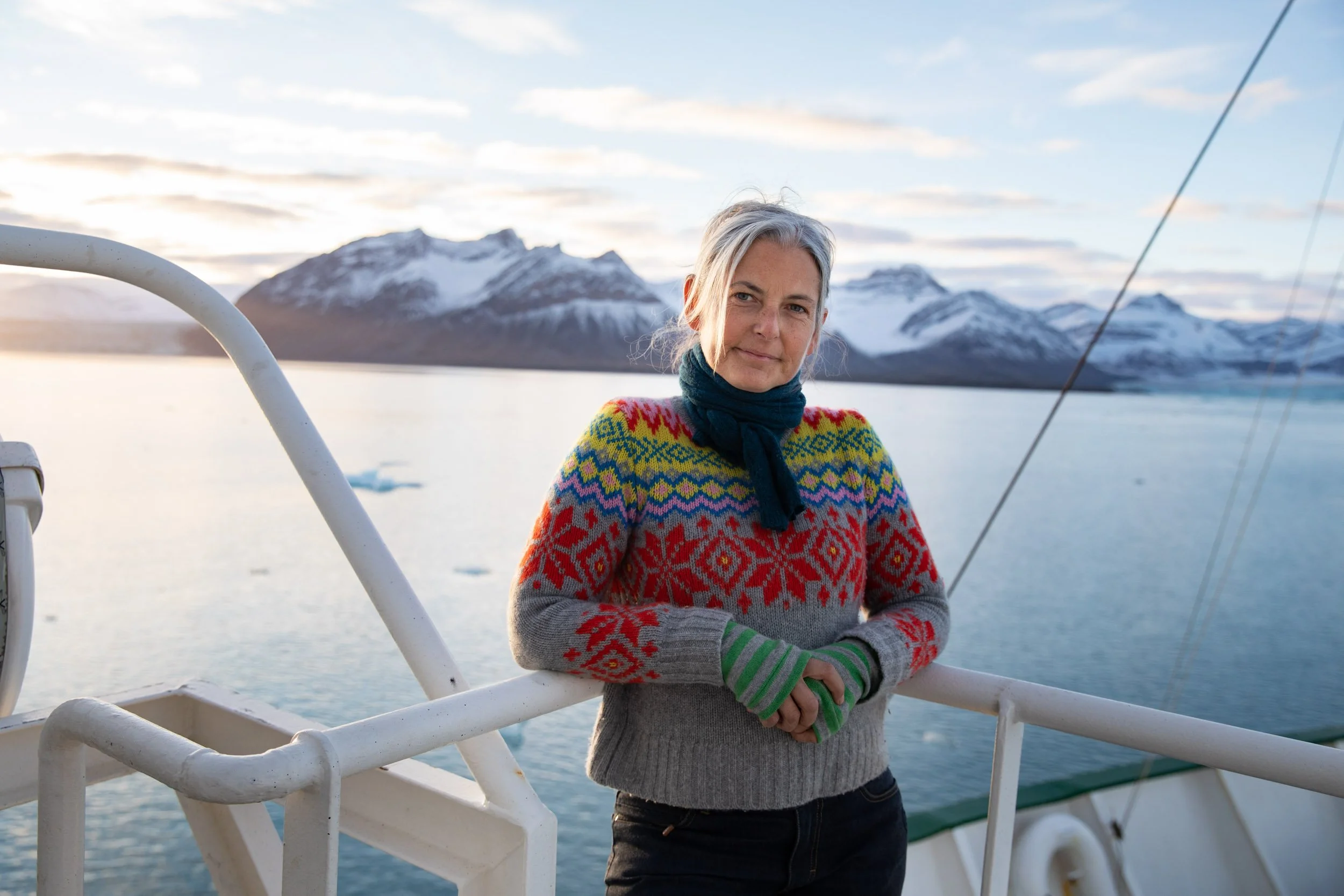 Woman in a colorful sweater standing on a boat with snow-covered mountains and icy water in the background