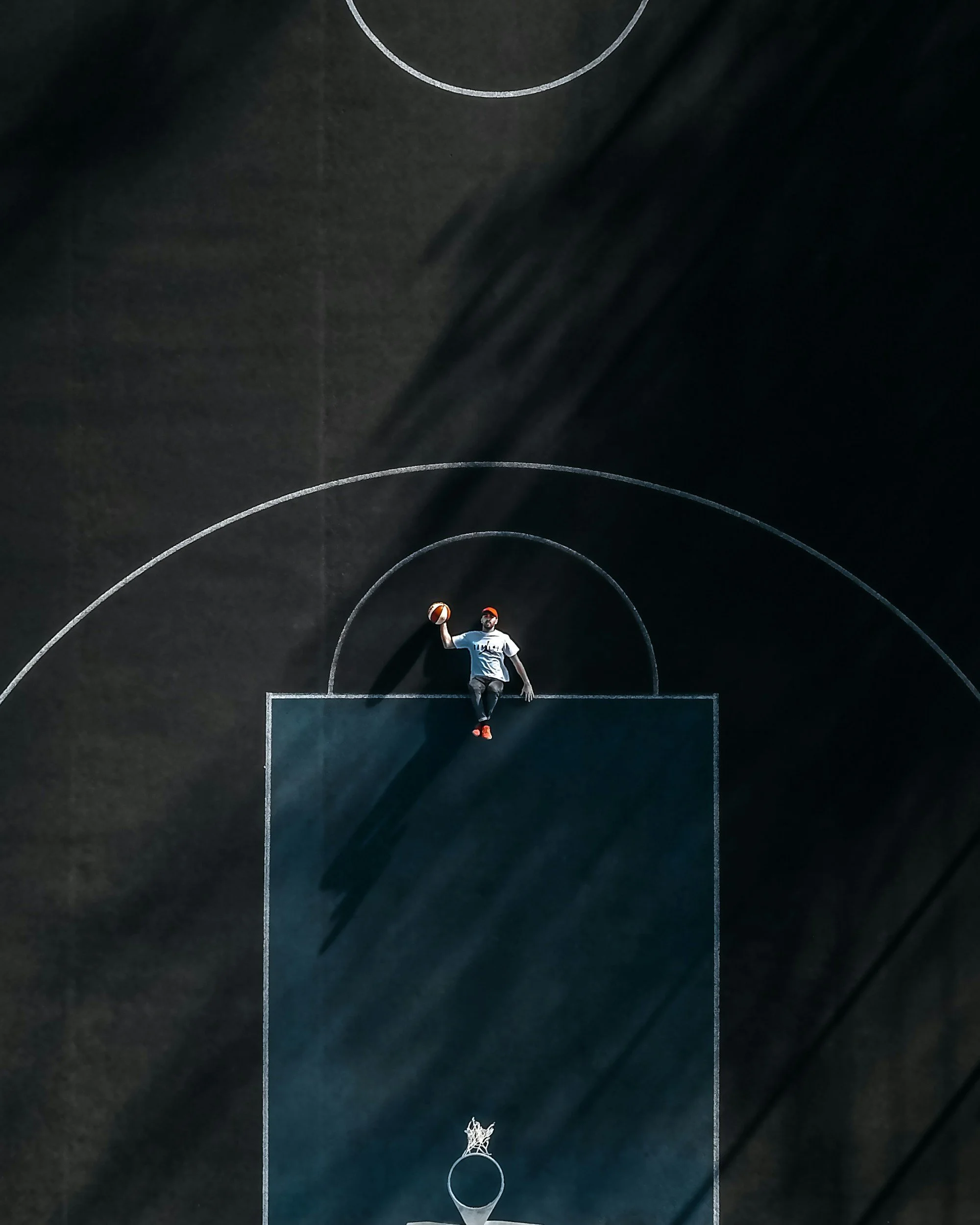 A person is sitting on a basketball court with one leg up and holding a basketball, seen from a drone camera shot of the court focusing on the court lines.