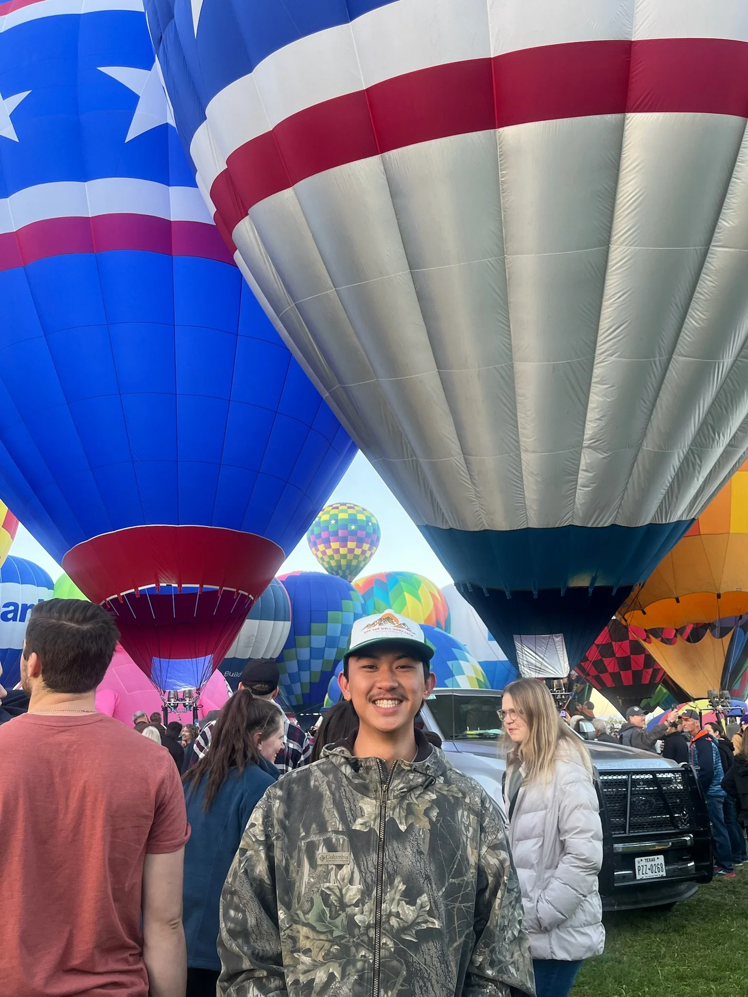 A man smiling at a hot air balloon festival with numerous colorful hot air balloons in the background, including one with the American flag design and a patriotic-themed balloon in the distance.