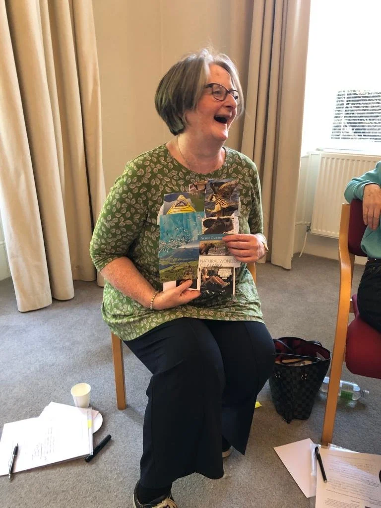 A woman with gray hair, glasses, and a green patterned shirt laughing and holding up several postcards or pictures, sitting on a wooden chair in a room with beige curtains and a window.