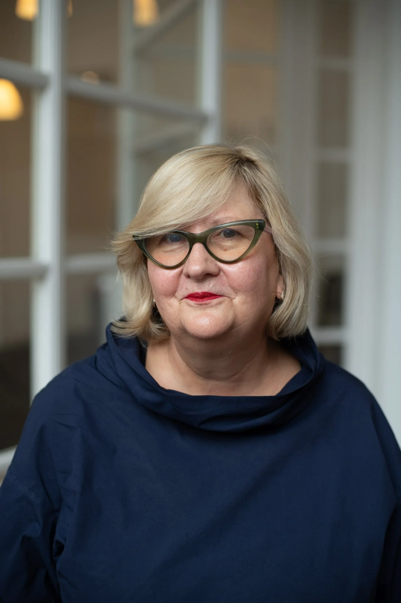 A middle-aged woman with blonde hair and glasses, wearing a dark blue top, standing indoors in front of a blurred background with white shelves.
