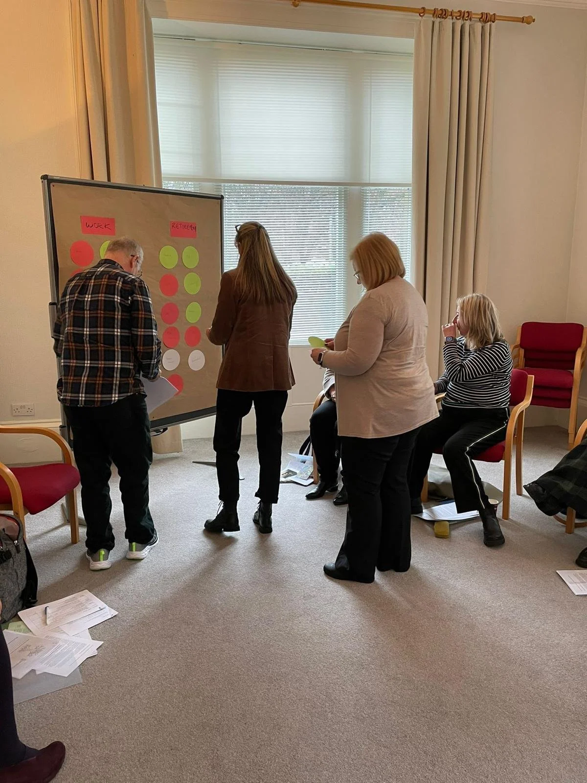 Group of five people participating in a workshop or discussion in a room with a large window and curtain. Two women are standing near a whiteboard with colored sticky notes, while three women are sitting, one taking notes and another observing.