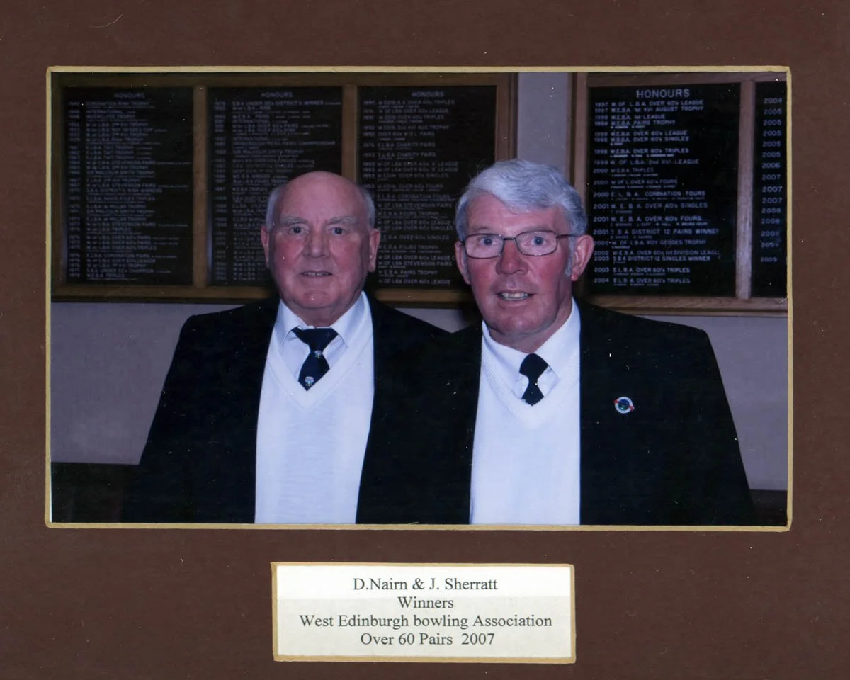 Two men dressed in formal attire, standing in front of a display board of awards, smiling at the camera. The plaque below indicates they are D. Nairn and J. Sherratt, winners of an over-60 pairs bowls competition in 2007 from the West Edinburgh Bowli