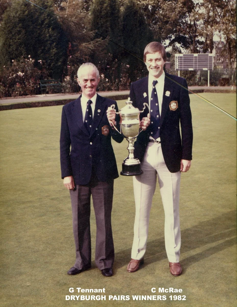 Two young men in golf uniforms standing on a golf course, holding a trophy together, celebrating their victory as 1982 Dryburgh Pairs Winners. They are smiling, wearing jackets with badges, and ties with polka dots.
