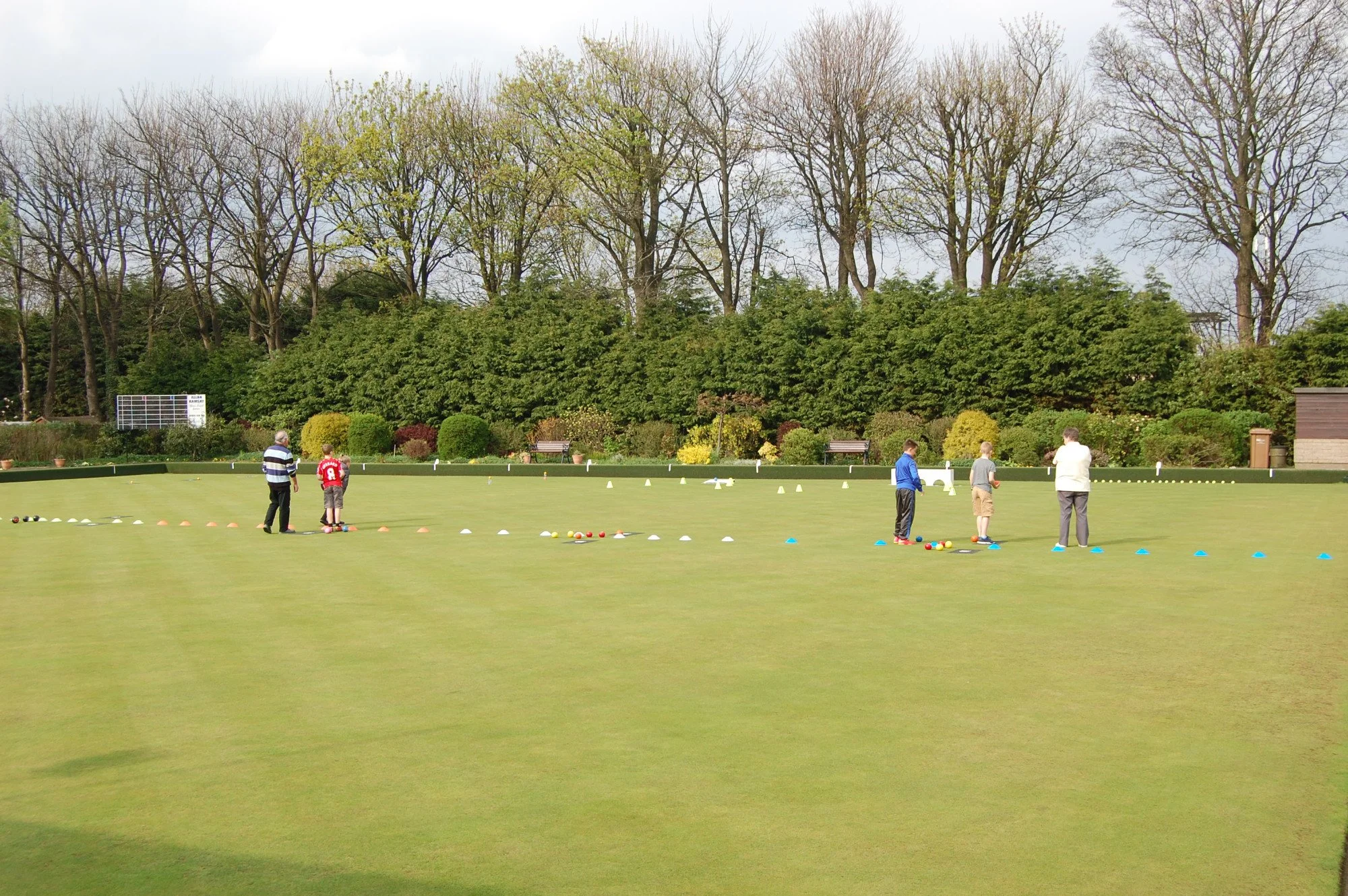 A group of children and an adult playing a game on a well-manicured lawn in a park, with cones, balls, and equipment set up for a sporting activity, and a backdrop of tall trees and bushes.