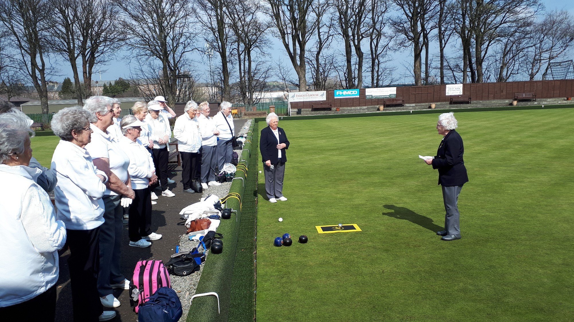A group of older women standing on the side of a lawn bowling green, listening to a woman who appears to be giving instructions or a speech, with lawn bowls placed on the ground nearby. The setting is outdoors with trees and benches in the background