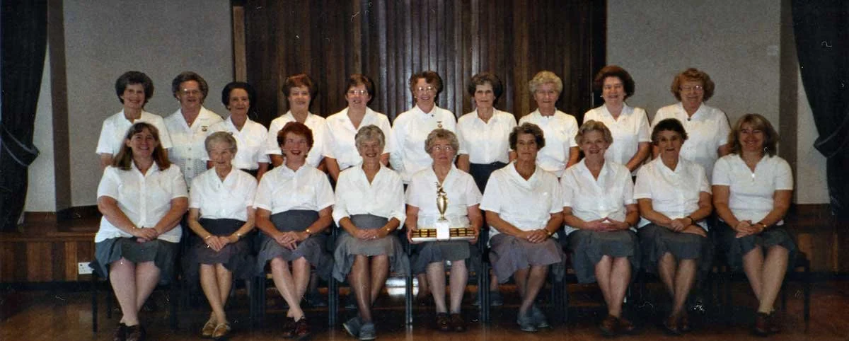 Group of women dressed in white blouses and dark skirts posing on a stage with curtains and a wood panel background, with a trophy placed in the center of the front row.