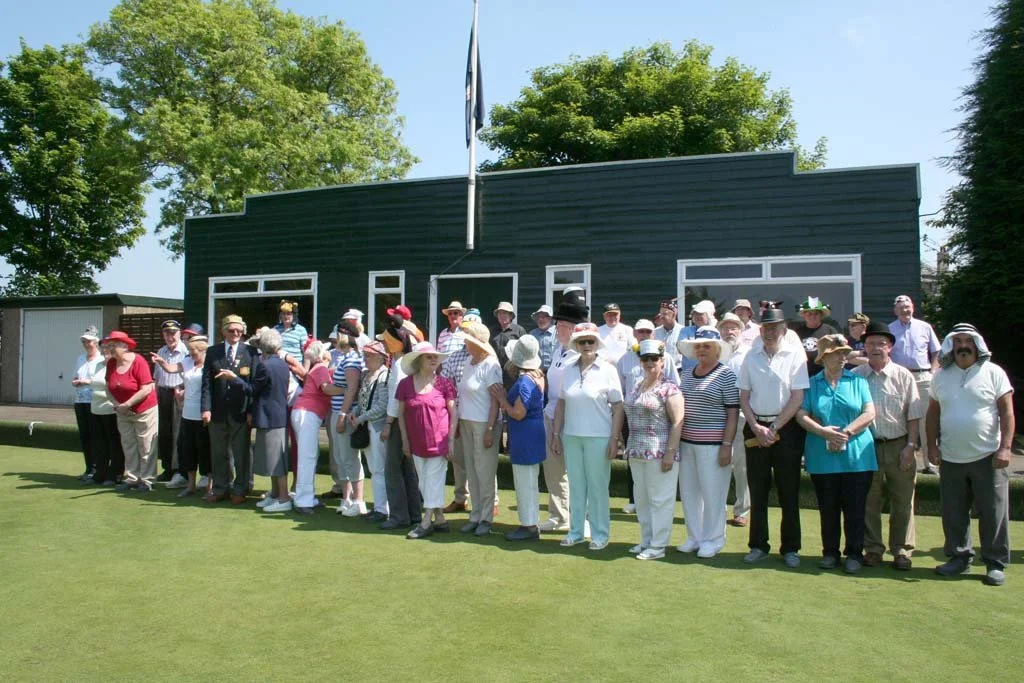 Group of older adults standing on a golf course, posing for a photo. They are dressed in casual and golf attire, with many wearing hats and sunglasses. Behind them is a dark building with large windows and a flagpole.