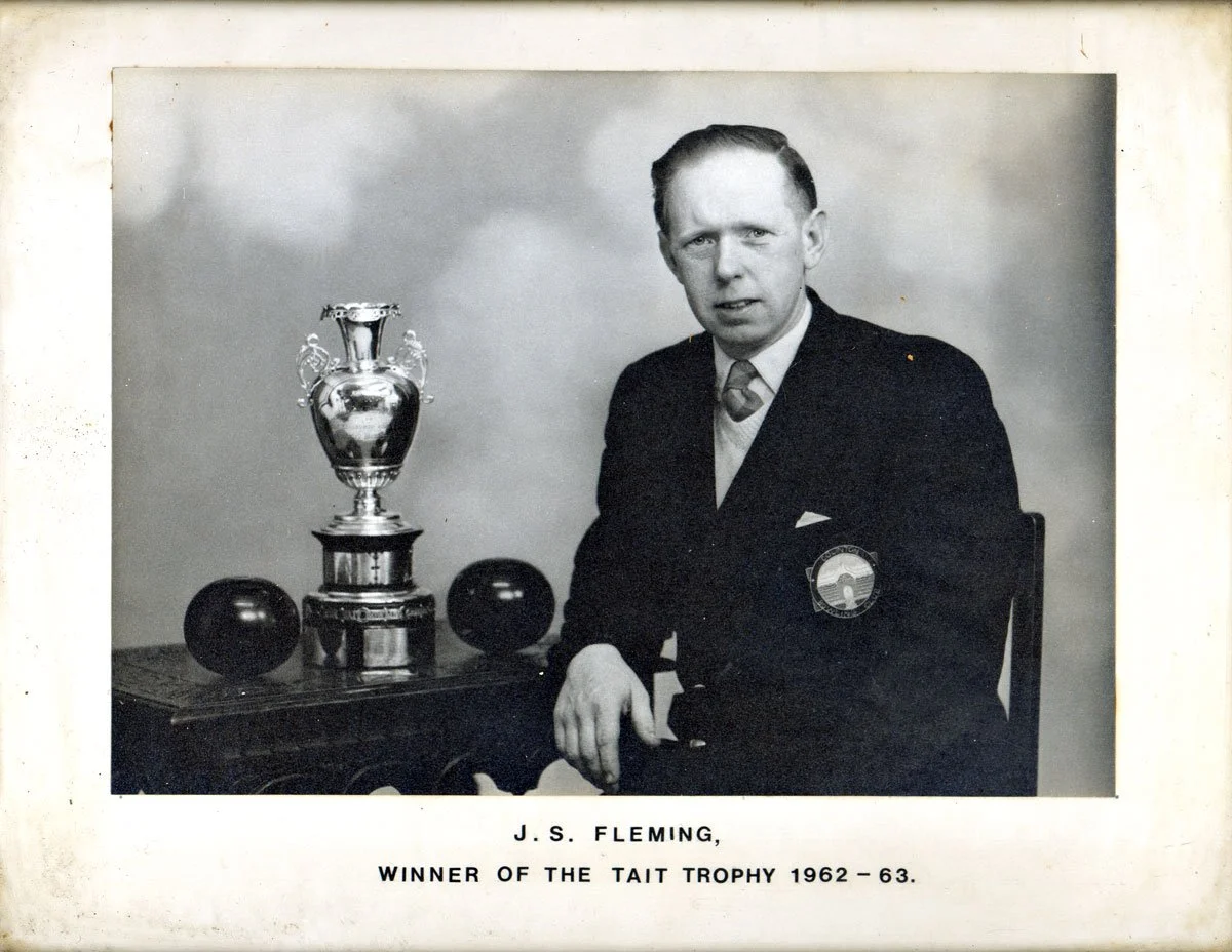 Black and white photograph of J.S. Fleming sitting next to a large trophy and two balls, with a plain background. The caption below identifies him as the winner of the Tait Trophy for 1962-63.
