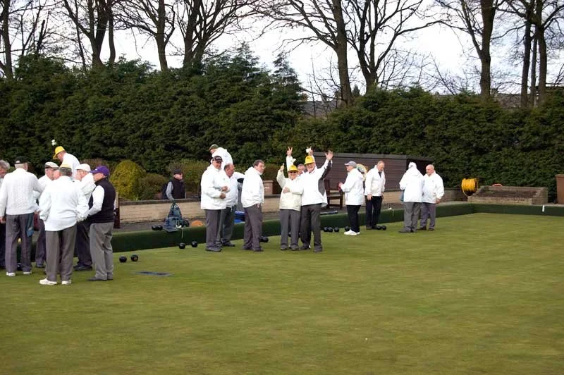 Group of people playing lawn bowling on a green, some wearing white clothing and yellow hats, others gathered around, with trees and shrubs in the background.