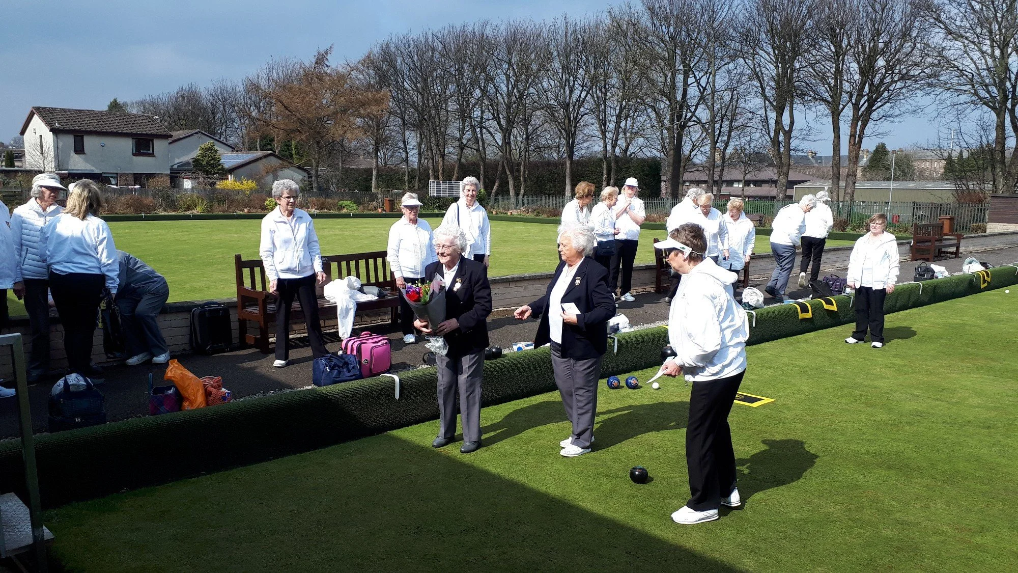 A group of elderly women gathering at a lawn bowling green on a sunny day, some wearing white jackets and hats, with one woman holding a flower bouquet, surrounded by bags and bowls.