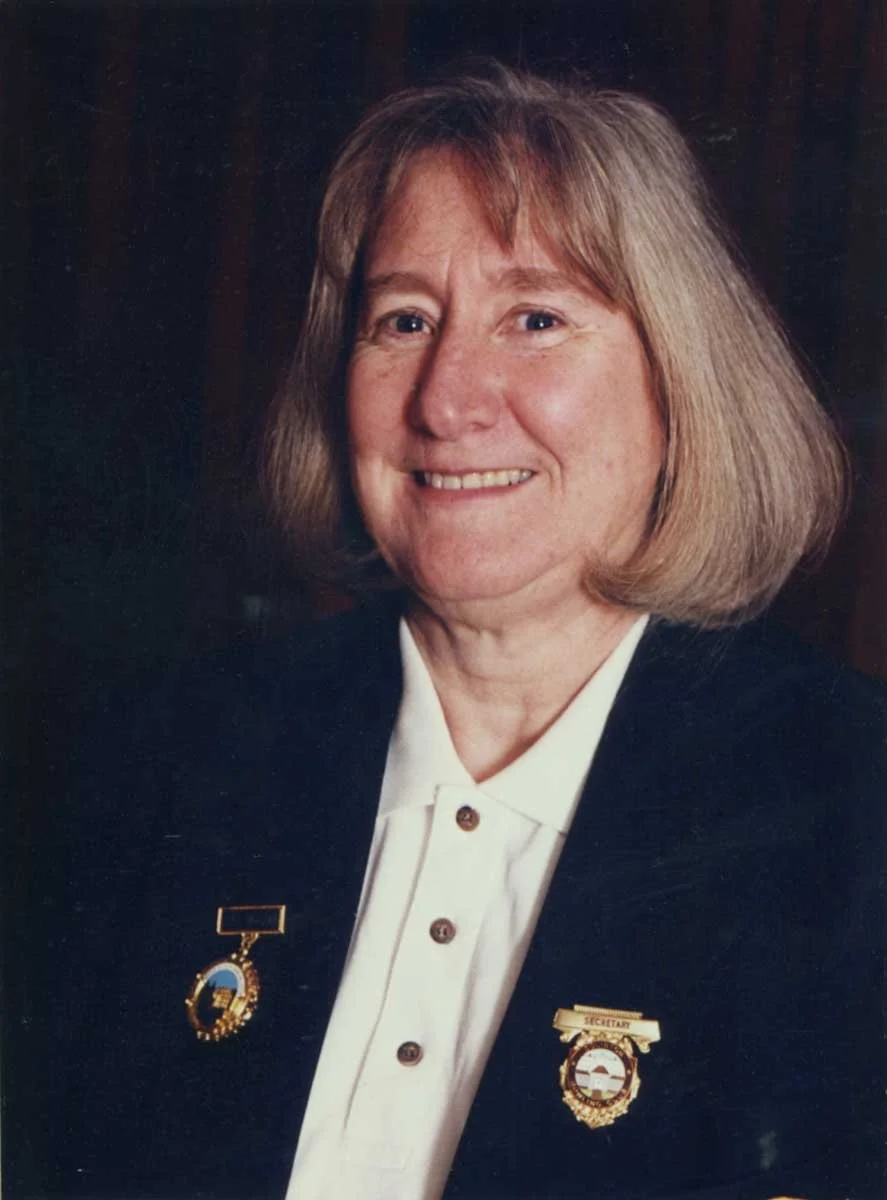 A middle-aged woman with shoulder-length blonde hair, wearing a black blazer with gold pins, a white button-up shirt, and smiling.