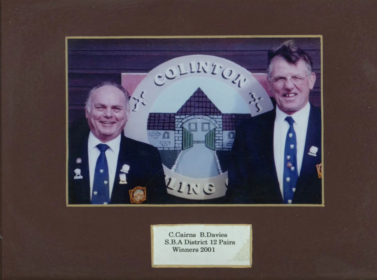 Photograph of two men in suits with badges, standing in front of a decorative sign that reads "Collinton Bowling" with a drawing of a house or building. Caption beneath the photo reads "C. Cairns B. Davies, S.B.A District 12 Pairs Winners 2001."
