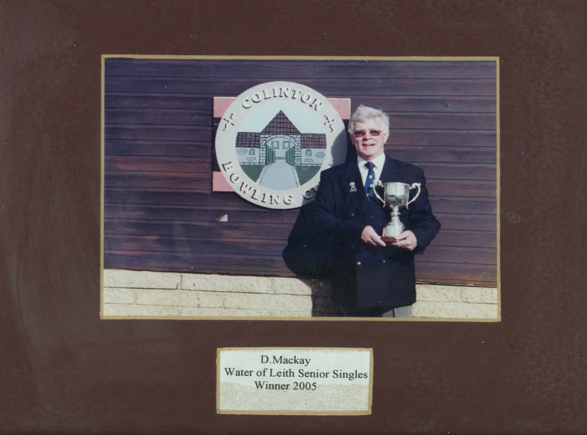 A man in a black suit with sunglasses holding a trophy in front of a wooden wall with a sign that reads 'Water of Leith Senior Singles Winner 2005'.