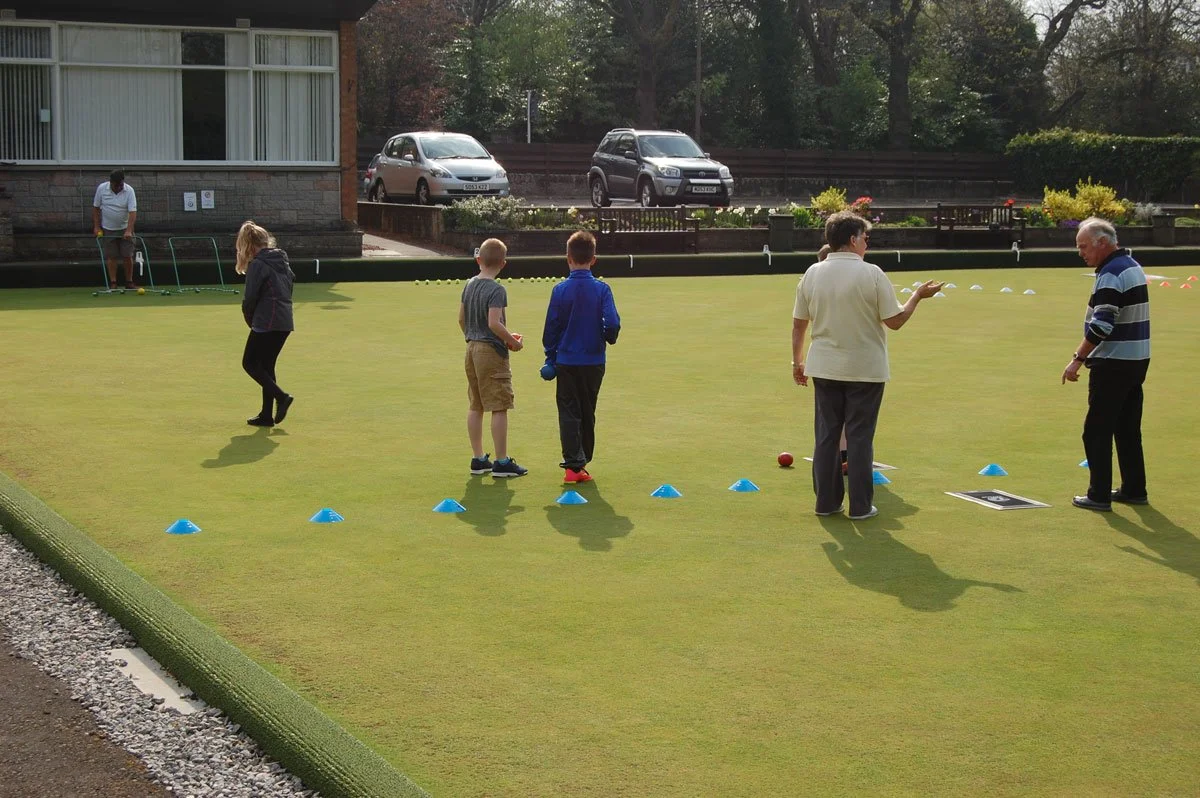 People practicing lawn bowling on a green, with cones set up and cars parked in the background.
