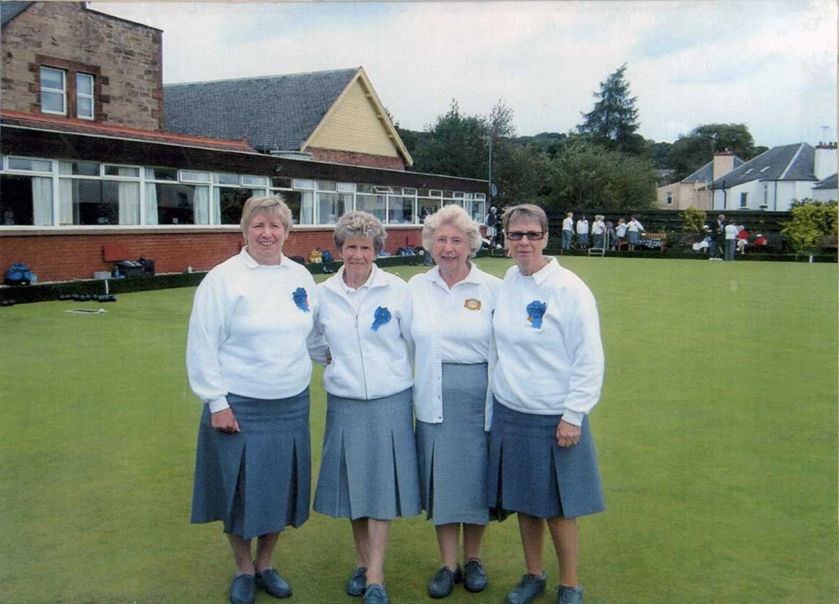 Four women in white sweaters with blue rosettes, standing on a lawn with a group of people in the background.
