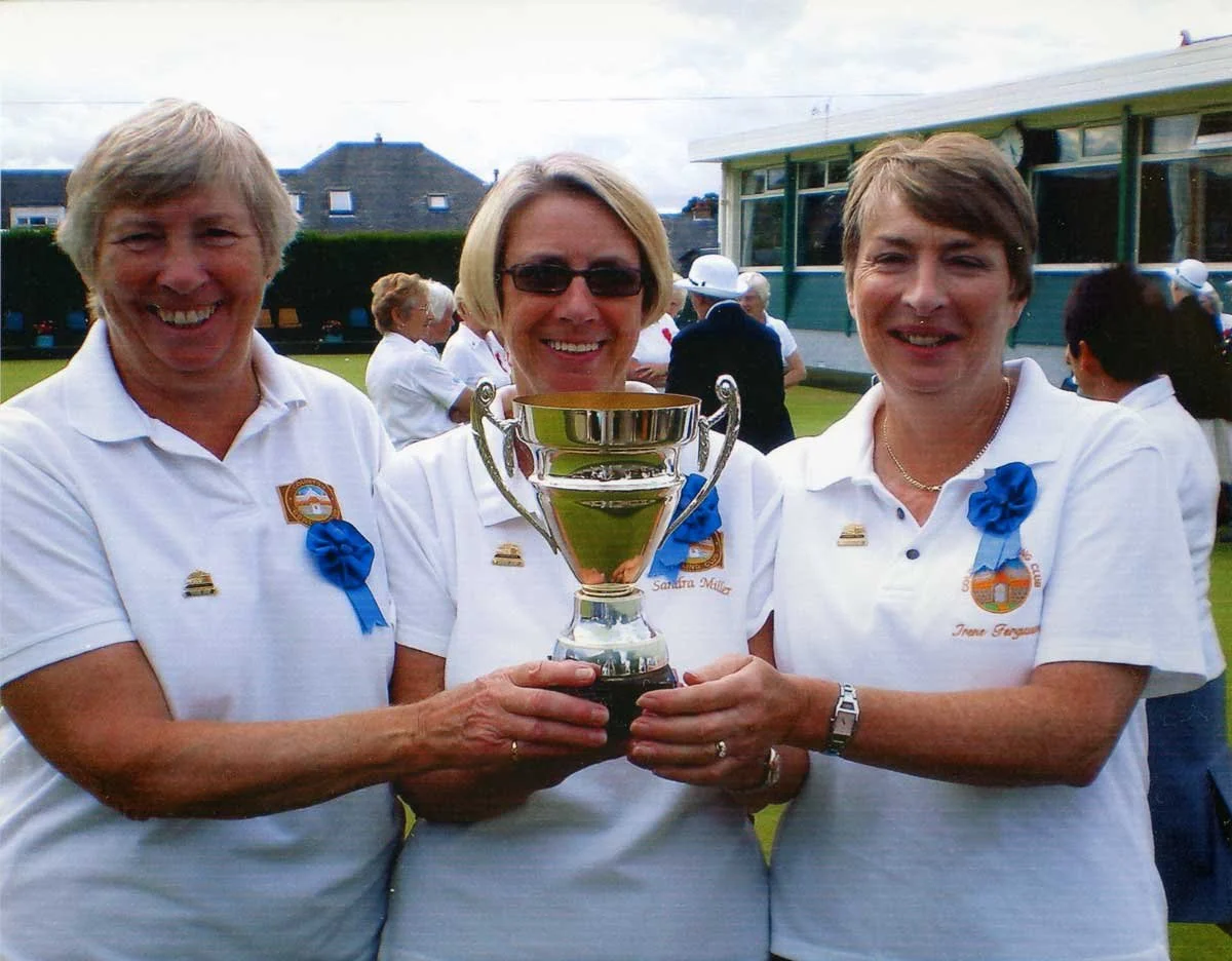 Three women in white shirts holding a trophy at a golf course, with other people and a clubhouse in the background.