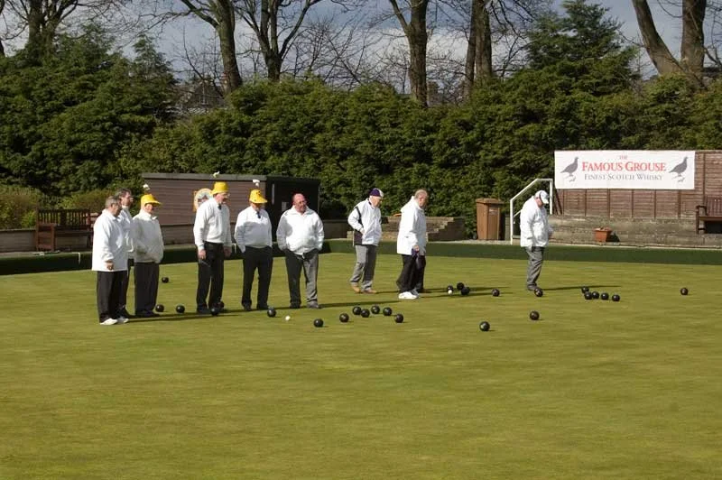 Group of people playing lawn bowls on a green field, some wearing yellow hats, with trees and a sign for a whiskey brand in the background.