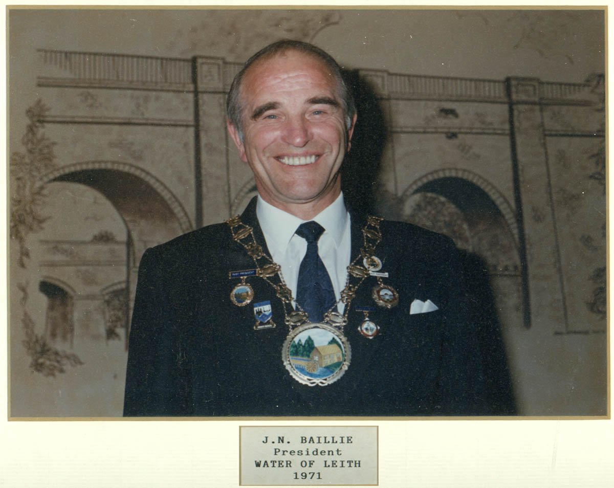 A man wearing a black suit with a chain of office and multiple pins, standing in front of a mural of a bridge. The plaque below identifies him as J.N. Baillie, President of Water of Leith in 1971.