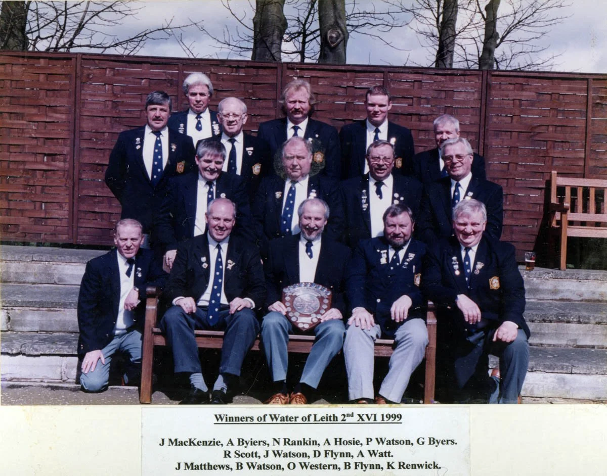 Group photo of 13 men dressed in dark blazers, white shirts, and ties, sitting and standing on outdoor steps with a wooden fence behind them. The men are holding or displaying plaques and medals. The photo is labeled as a 1999 reunion of water winner